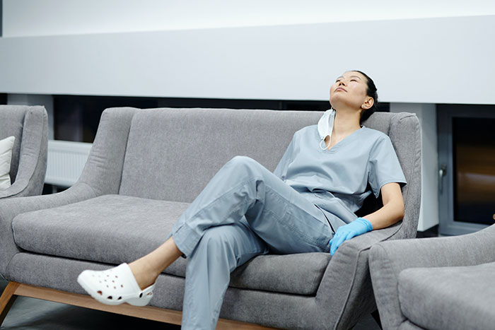 Woman welcomes brother’s family for a month, sitting exhausted on a sofa wearing scrubs and gloves at home. Woman welcomes brother’s family for a month, sitting exhausted on a sofa wearing scrubs and gloves at home.