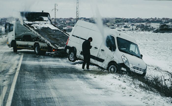 White van stuck in icy roadside ditch with tow truck and person assessing the situation, showcasing instant karma example.