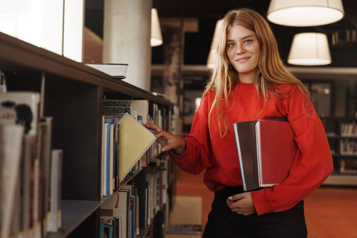 Young woman in a red sweater selecting a book from a library shelf, illustrating small decisions with unexpected consequences.