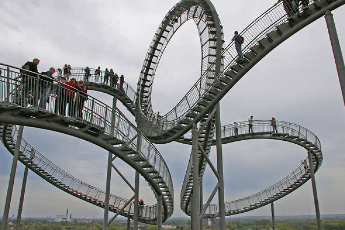 Looping pedestrian bridge with people walking, one appearing to walk a vertical loop, in places that look AI generated but are real.