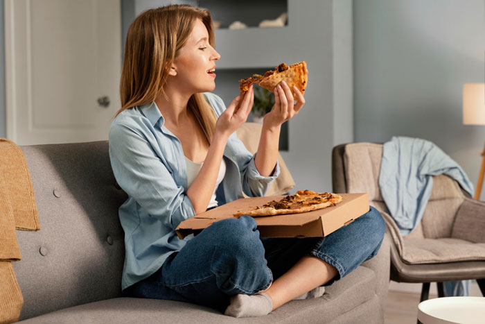 Woman sitting on a couch enjoying a slice of pizza, illustrating lazy relatives not bringing food to Christmas dinner debate.