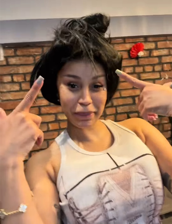 Cardi B pointing to new neck and back piercings with a playful expression against a brick wall background.