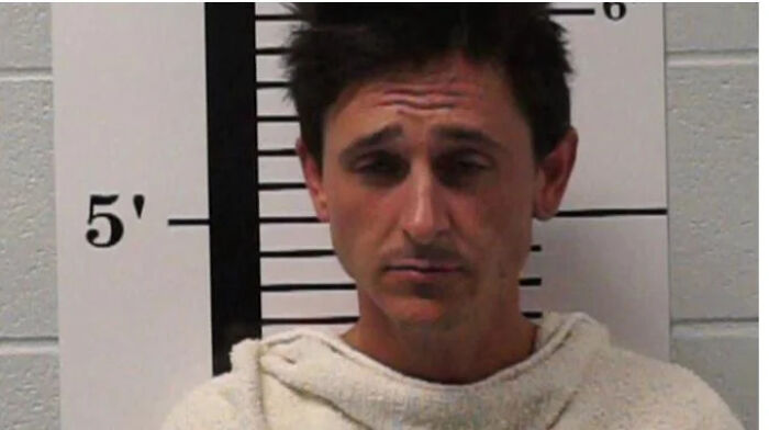 Man in a white shirt standing against height chart in a famous celebrity mugshot for reasons they got arrested