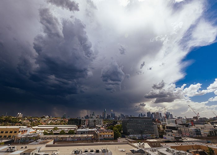 Dark chaotic storm clouds over a city skyline contrasting with bright blue sky, creating an intense dramatic scene.