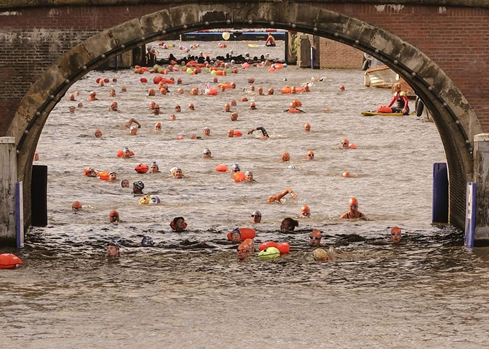 Crowded chaotic scene of swimmers in a river passing under a stone bridge during an open water event.