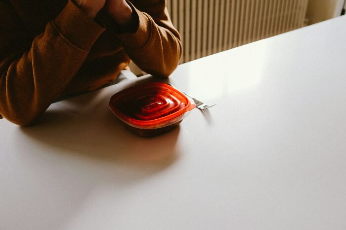 Person in a brown sweater sitting alone at a table with a sealed food container, reflecting on workplace scandals.