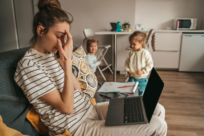 Stressed aunt ignoring unruly niblings playing in background with laptop on her lap in a cozy living room. Stressed aunt ignoring unruly niblings playing in background with laptop on her lap in a cozy living room.