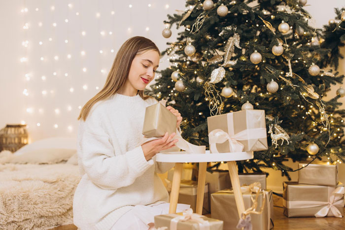 Woman in white sweater wrapping Christmas gifts by decorated tree, illustrating aunt no Christmas gifts for unruly niblings. Woman in white sweater wrapping Christmas gifts by decorated tree, illustrating aunt no Christmas gifts for unruly niblings.