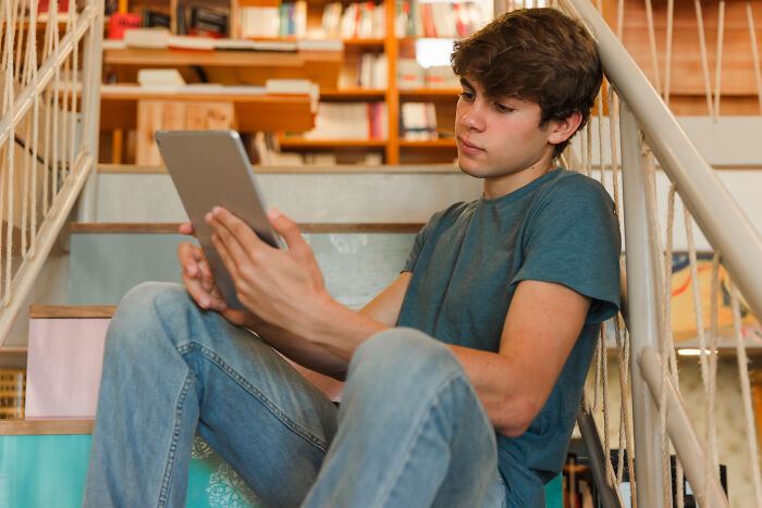 Teen boy sitting on stairs reading a tablet, looking thoughtful with bookshelves behind, related to Christmas nanny kid bite. Teen boy sitting on stairs reading a tablet, looking thoughtful with bookshelves behind, related to Christmas nanny kid bite.