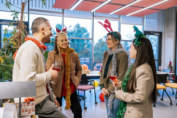 Group of coworkers enjoying a festive work Christmas party, wearing holiday accessories and holding drinks indoors.
