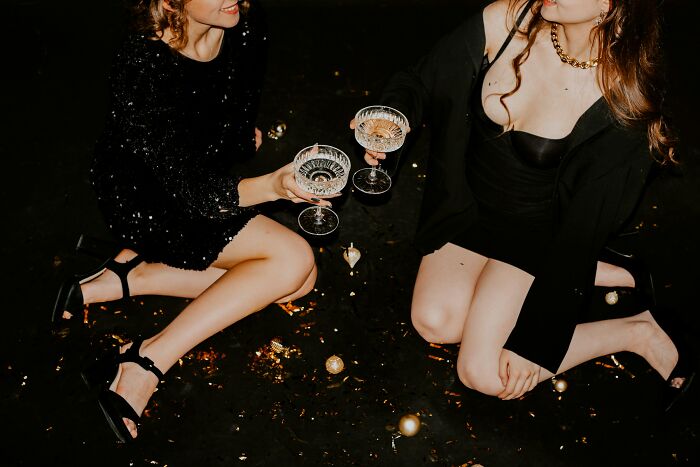 Two women in elegant black dresses sitting on the floor, clinking glasses at a wild work Christmas party celebration.