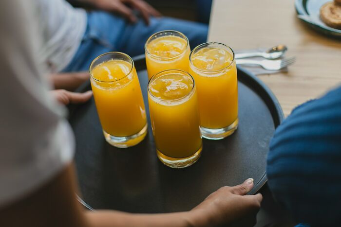 Four glasses of orange juice on a tray being served at a work Christmas party with people gathered around.