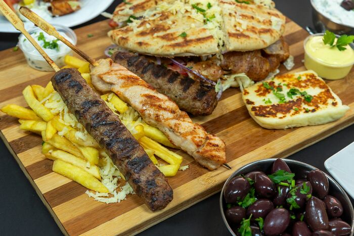 Grilled Greek skewers, fries, pita bread, olives, and cheese served on a wooden platter in a Greek village meal setting Grilled Greek skewers, fries, pita bread, olives, and cheese served on a wooden platter in a Greek village meal setting