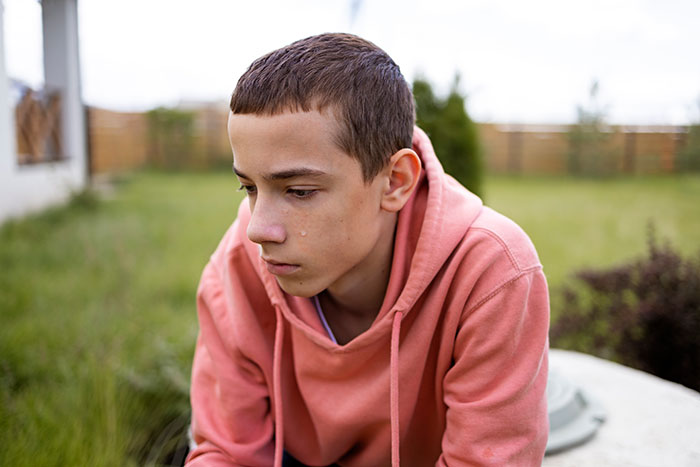 Exhausted teen in pink hoodie sitting outdoors, tear on cheek, looking sleepless and distressed