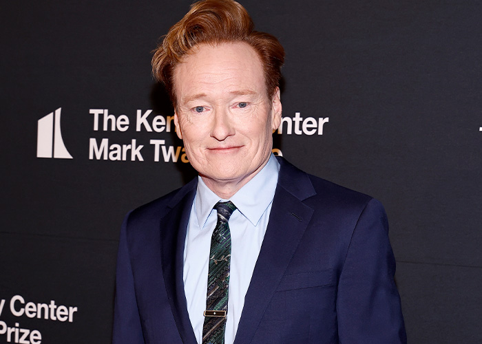 Conan O&rsquo;Brien wearing a navy suit and patterned tie at a media event with a dark backdrop and white text.