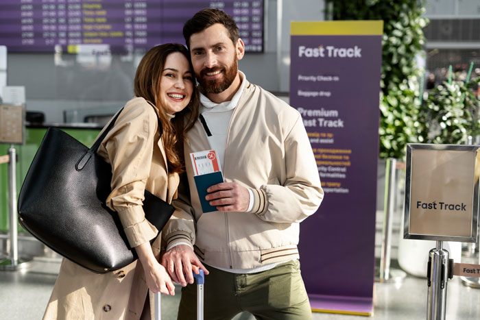 Happy couple at airport check-in counter near fast track sign holding boarding passes and luggage.