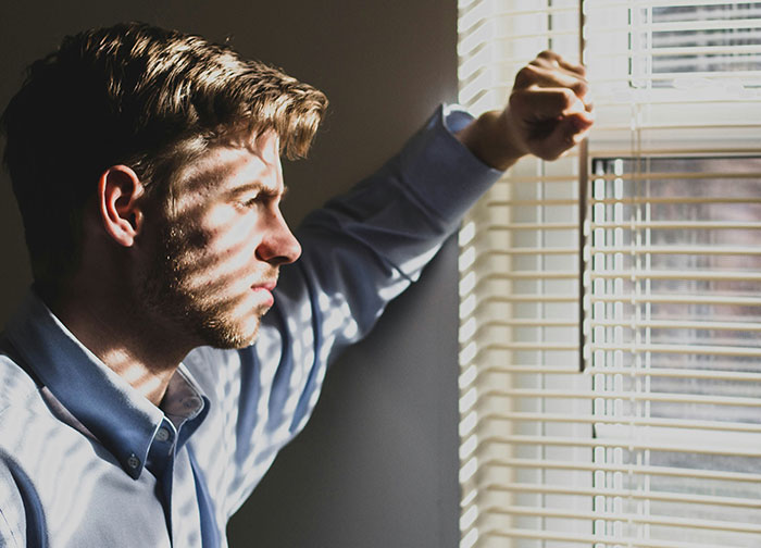 Young man looking out window blinds with a serious expression, reflecting on relationship conflicts about closing closet door. Young man looking out window blinds with a serious expression, reflecting on relationship conflicts about closing closet door.