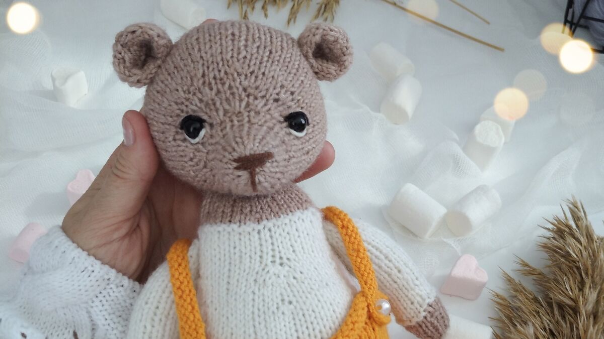 Hand holding a handmade teddy bear knitted from a ball of yarn with white and orange clothing details.