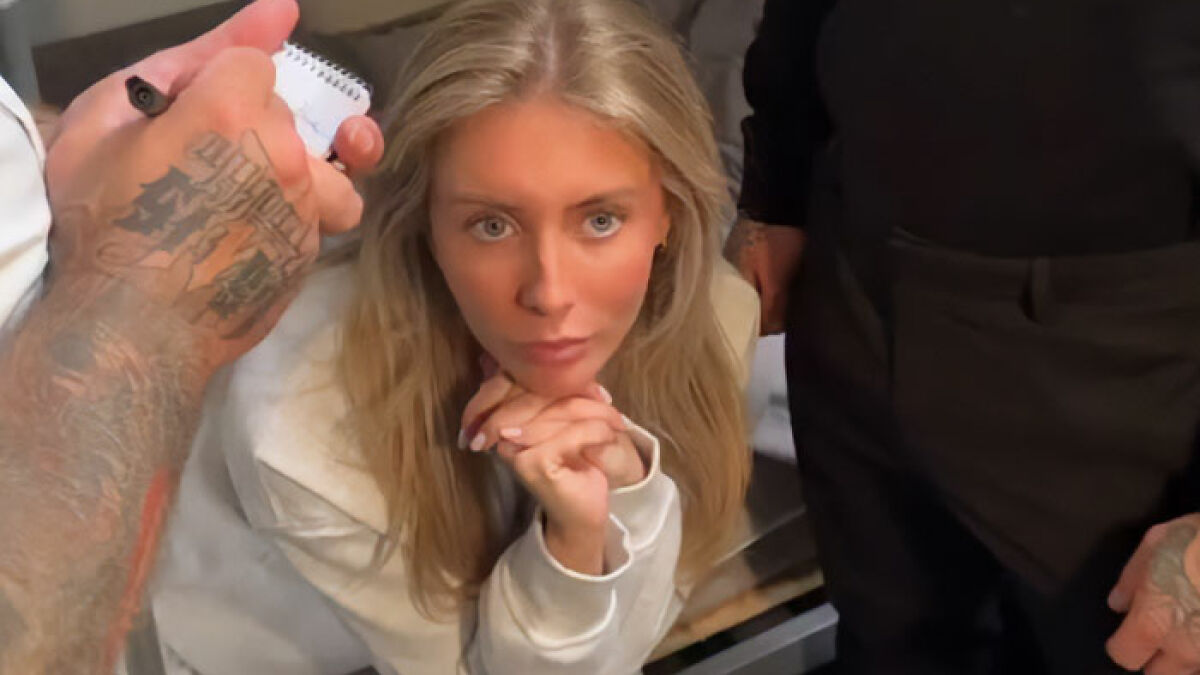 Blonde woman with defiant expression sitting and resting chin on hands in a dimly lit room with tattooed arms nearby
