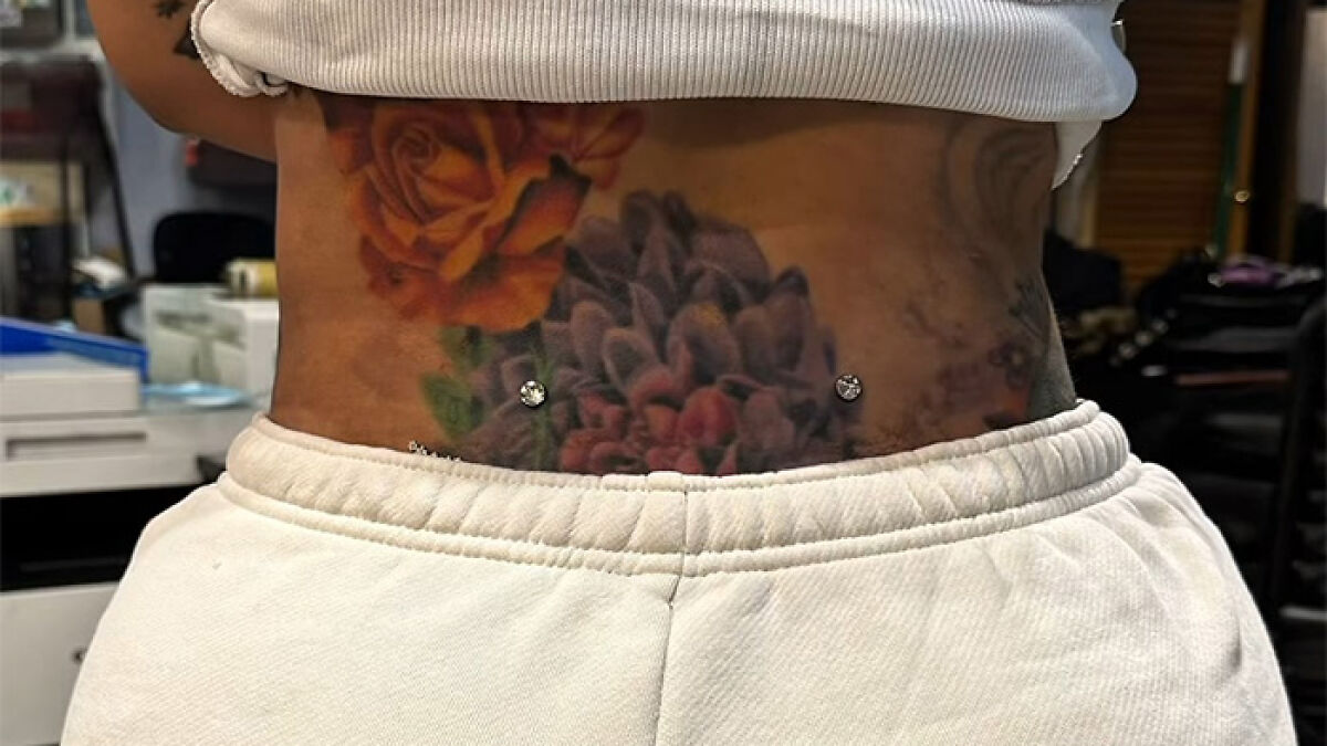 Cardi B showing new neck and back piercings above colorful floral tattoos, wearing white pants and a cropped top.