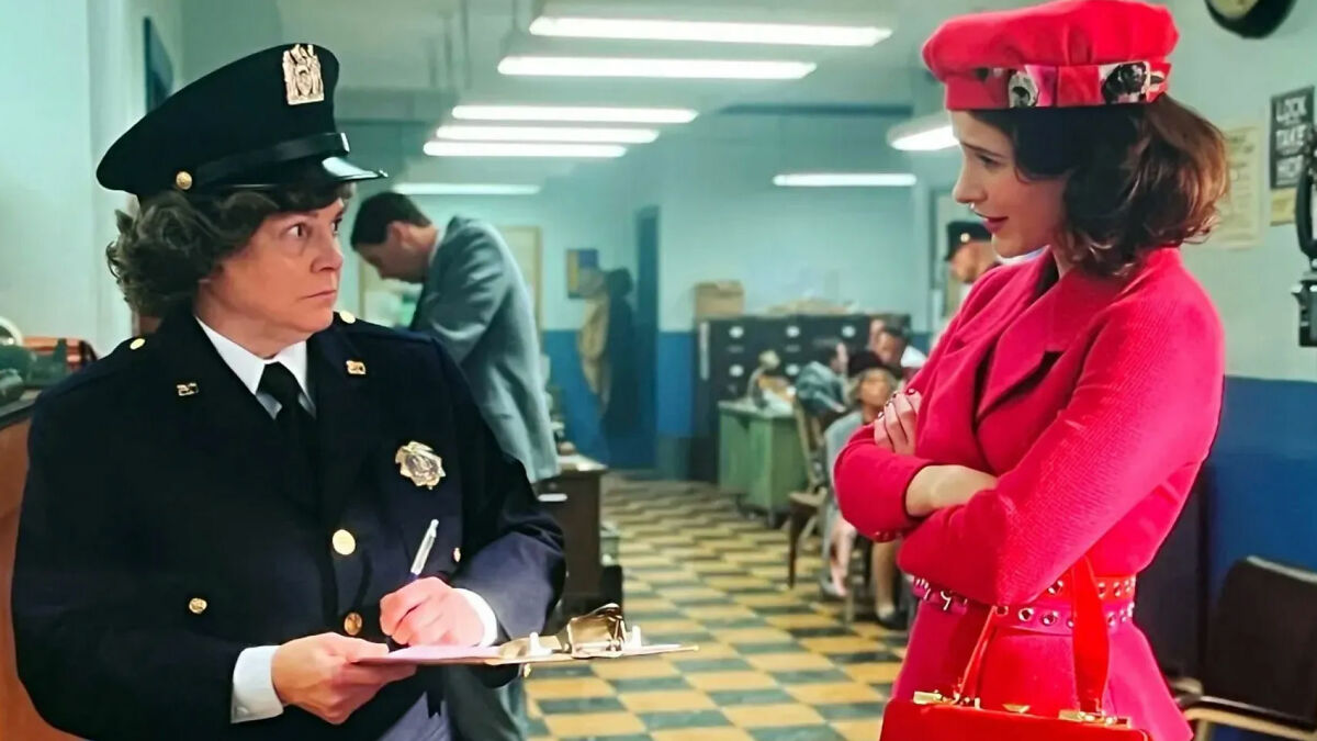 Actress from Marvelous Mrs. Maisel in red outfit talking to a police officer inside a retro police station.