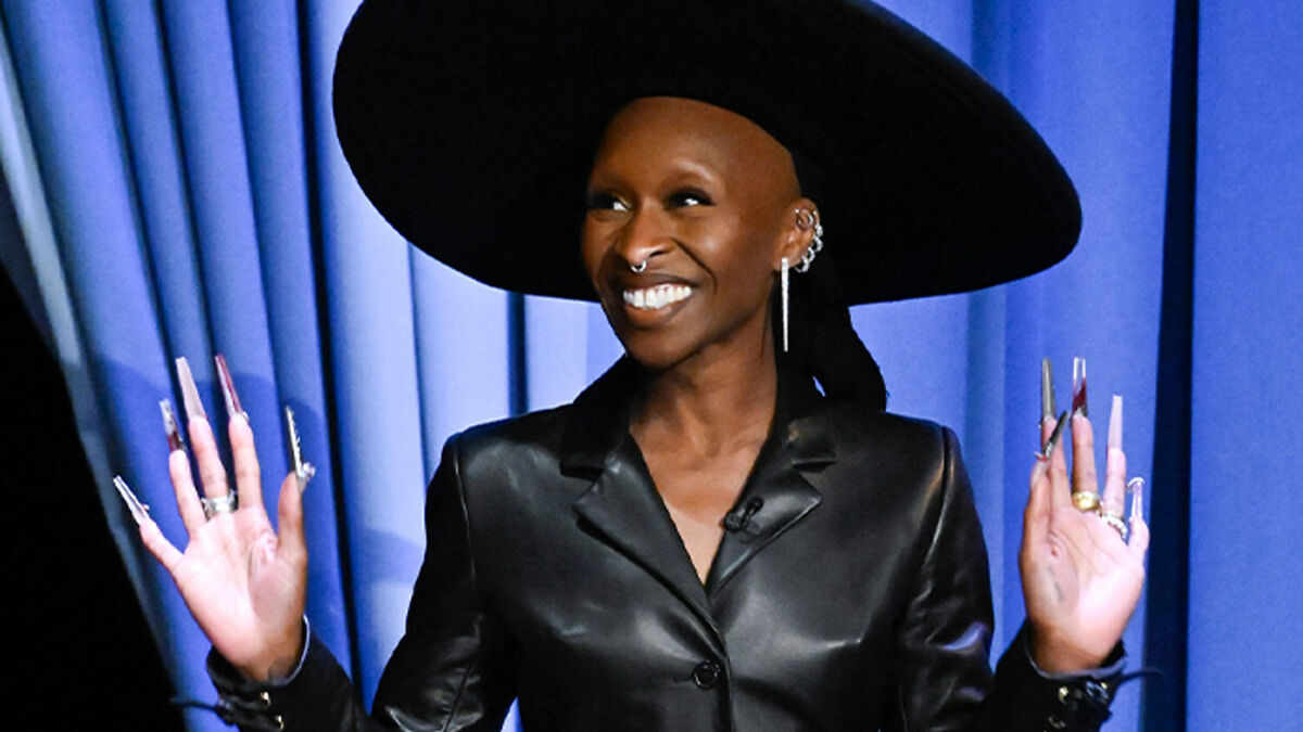 Cynthia Erivo smiling in a black outfit and wide hat, showing her oddly large hands with long decorated nails.