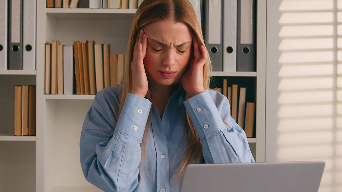 Frustrated coworker holding her head in office with laptop, stressed about fixing her own mistake at work.