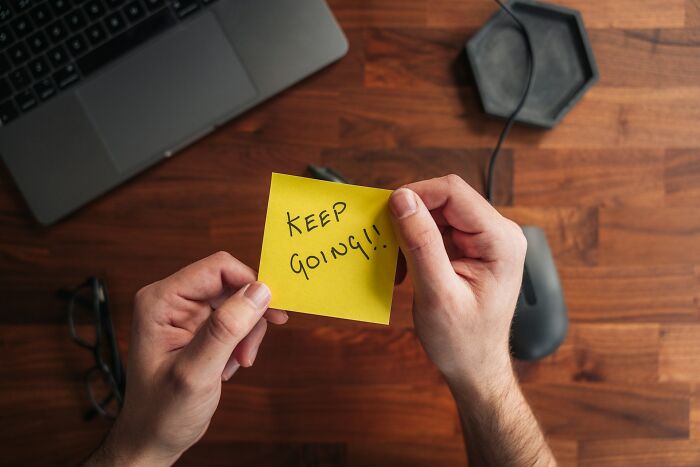 Hands holding a sticky note with keep going written, a laptop and glasses on a wooden desk, showing harmful but toxic habits.