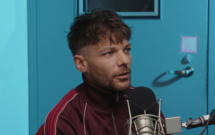 Man in a maroon jacket speaking into microphone during a podcast about running into different celebrities.