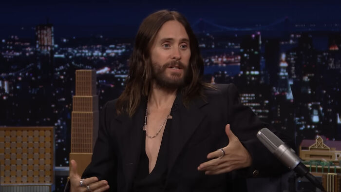 Actor Jared Leto in a black blazer speaking on a late night talk show with city lights in the background, celebrity encounter.
