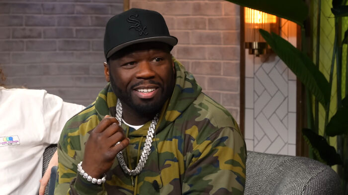 Man in camo hoodie and black cap smiling during casual celebrity encounter on a talk show set with modern decor.