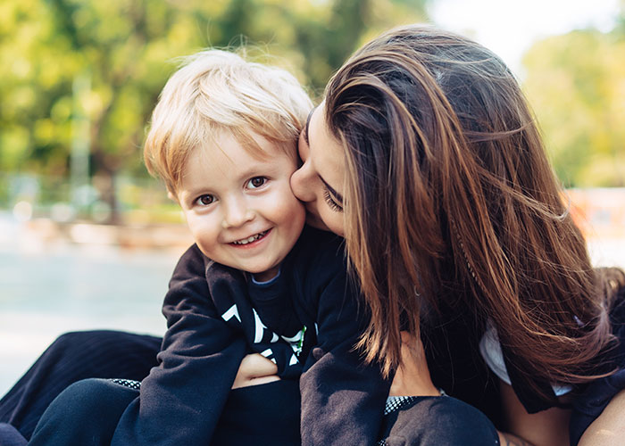 Young woman hugging and kissing smiling blonde toddler boy outdoors on a sunny day, family dog incident discussed.