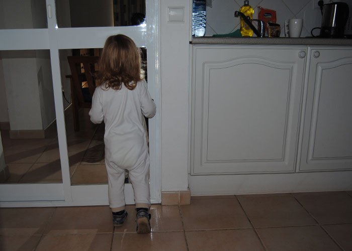 Toddler in white pajamas standing by a glass door in a dimly lit room, evoking creepy things kids said moments.