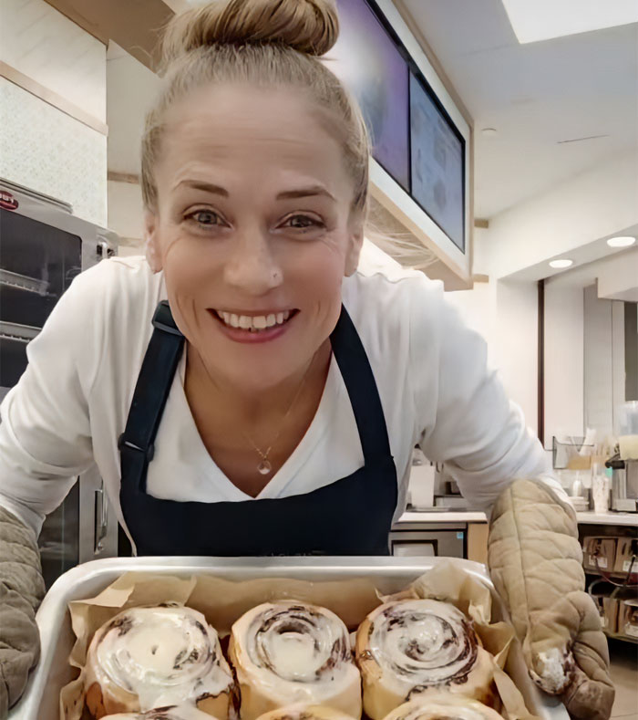 Cinnabon worker smiling behind a tray of cinnamon rolls in a bakery setting, linked to viral racist tirade controversy. Cinnabon worker smiling behind a tray of cinnamon rolls in a bakery setting, linked to viral racist tirade controversy.