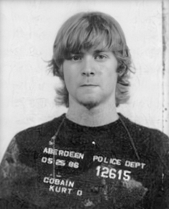 Black and white infamous celebrity mugshot of a young man with medium-length hair holding a police ID board.