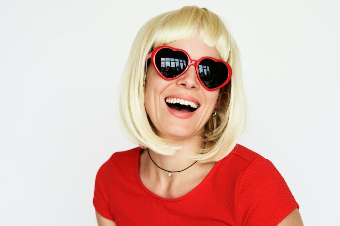 Woman with blonde bob haircut wearing red heart-shaped sunglasses and a red shirt, smiling in a casual setting for toxic habits.