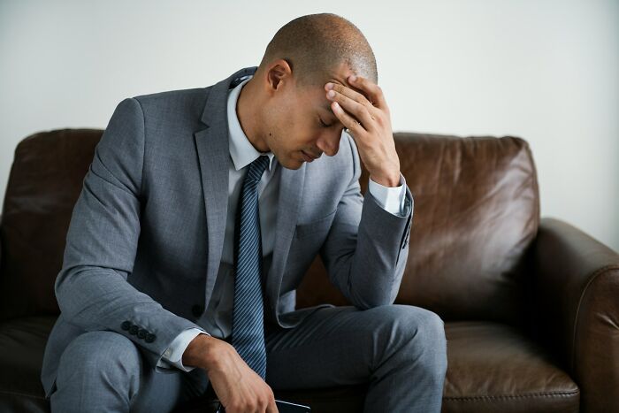 Man in gray suit sitting on brown leather couch, holding head in hand, stressed by crazy airport stories.