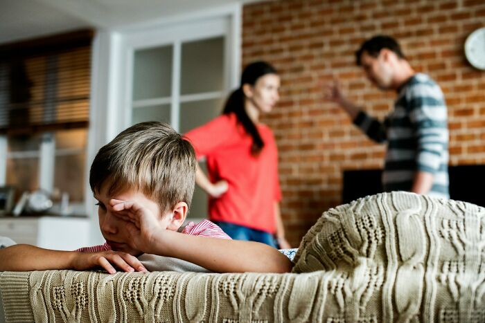 Young boy wiping tears while parents argue in background, highlighting challenges of modern parenting trends.