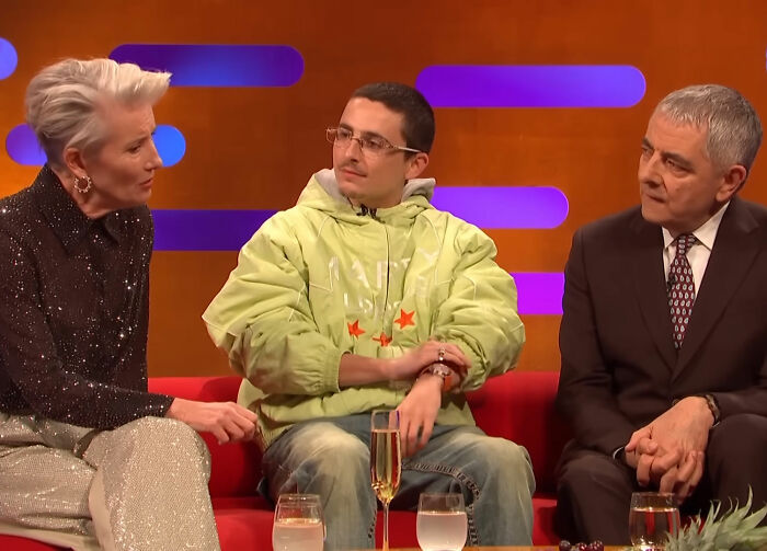 Timothée Chalamet on a talk show couch, wearing glasses and a green hoodie, with guests in conversation. Timothée Chalamet on a talk show couch, wearing glasses and a green hoodie, with guests in conversation.
