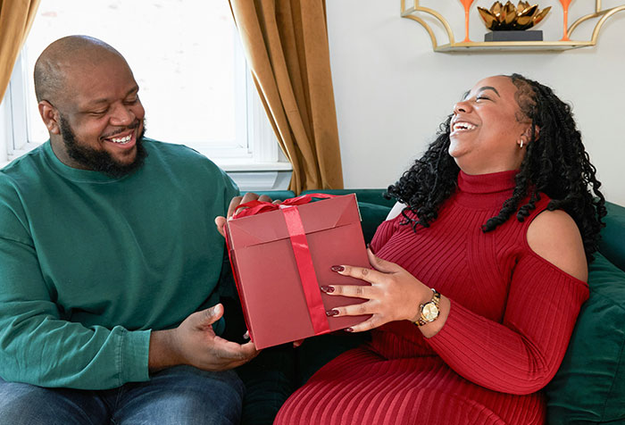 Man and woman laughing as woman hands man a red Christmas gift box, capturing a moment of petty Christmas revenge.