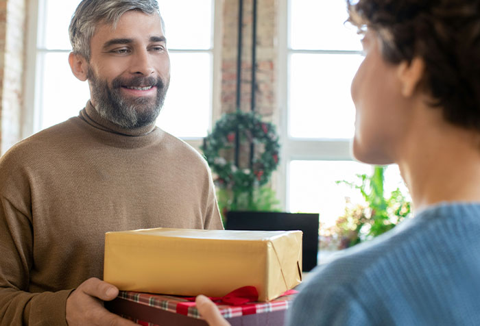 Man smiling while giving wrapped Christmas gifts to another person, capturing petty Christmas revenge moment.