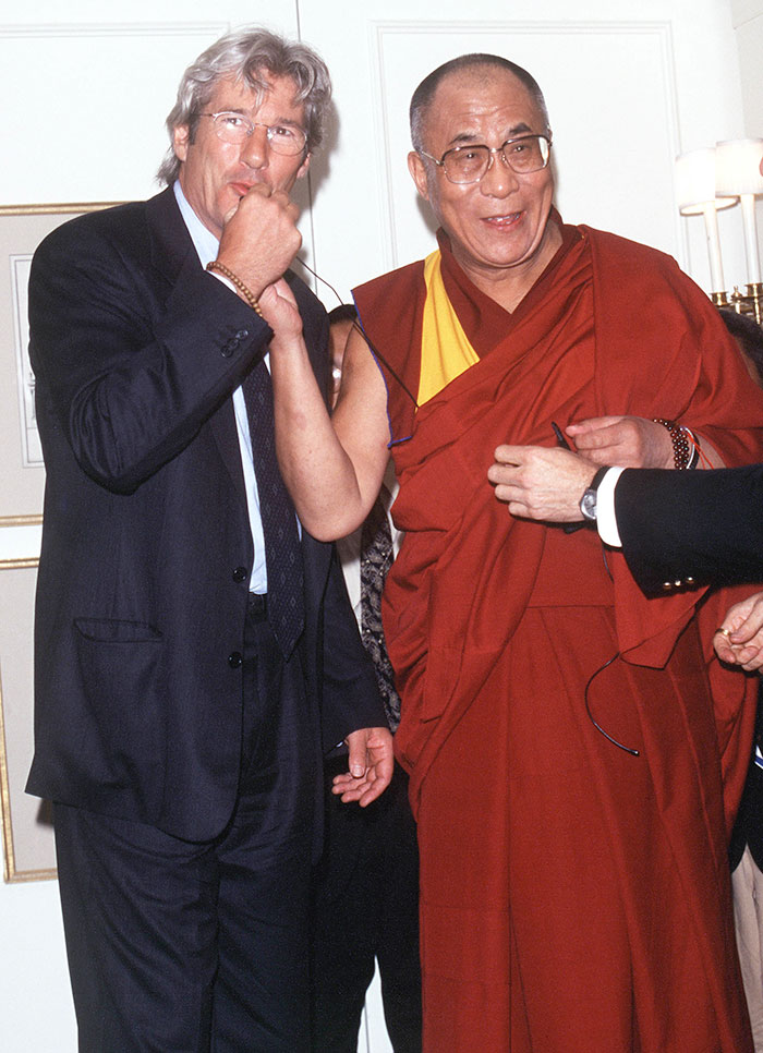 Richard Gere in a suit standing next to a man in traditional red and yellow robes during a casual event. Richard Gere in a suit standing next to a man in traditional red and yellow robes during a casual event.