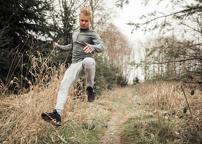 Young man jumping over a narrow trail in a forest, illustrating survival hacks that can be dangerous and useless.