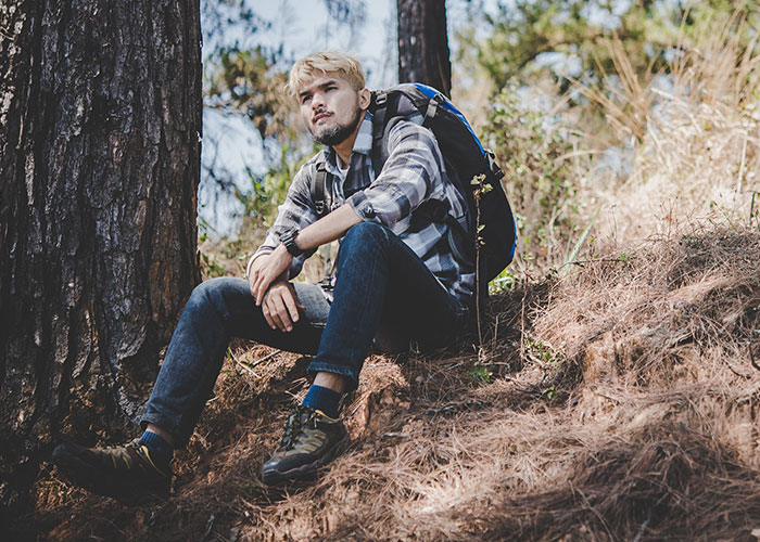 Young man with backpack sitting in the forest looking thoughtful, illustrating survival hacks that can be dangerous.
