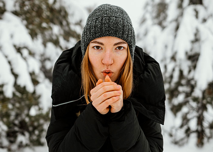 Young woman in winter clothing warming her hands in a snowy forest, illustrating survival hacks in cold environments.