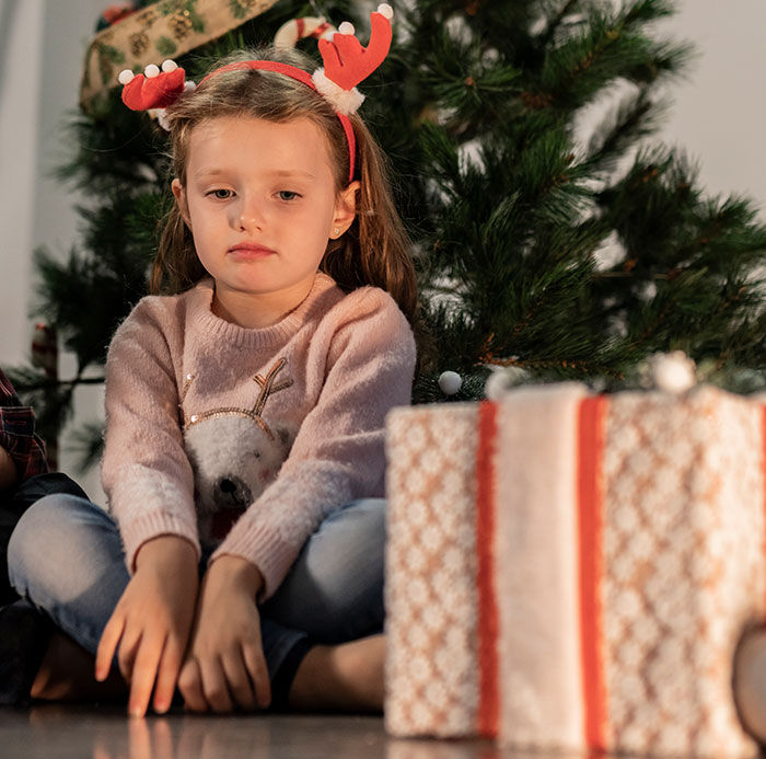 Sad 7-year-old girl with reindeer antlers sitting by Christmas tree and unopened gifts, reflecting holiday punishment debate.