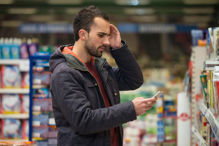 Man reading phone with concerned expression in store aisle, reflecting stress from caring for autistic brother situation. Man reading phone with concerned expression in store aisle, reflecting stress from caring for autistic brother situation.