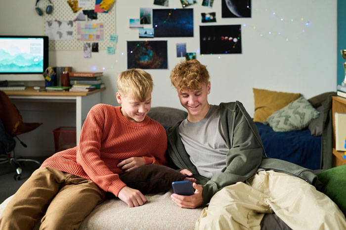 Two teenage boys sitting on a bed smiling at a smartphone, depicting teen mad as dad gifts stepbro a car for Xmas. Two teenage boys sitting on a bed smiling at a smartphone, depicting teen mad as dad gifts stepbro a car for Xmas.