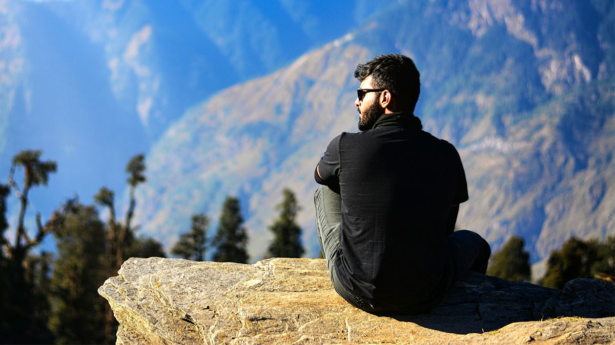Man in sunglasses sitting on a rock overlooking mountains, representing the digital nomad lifestyle and remote work freedom.