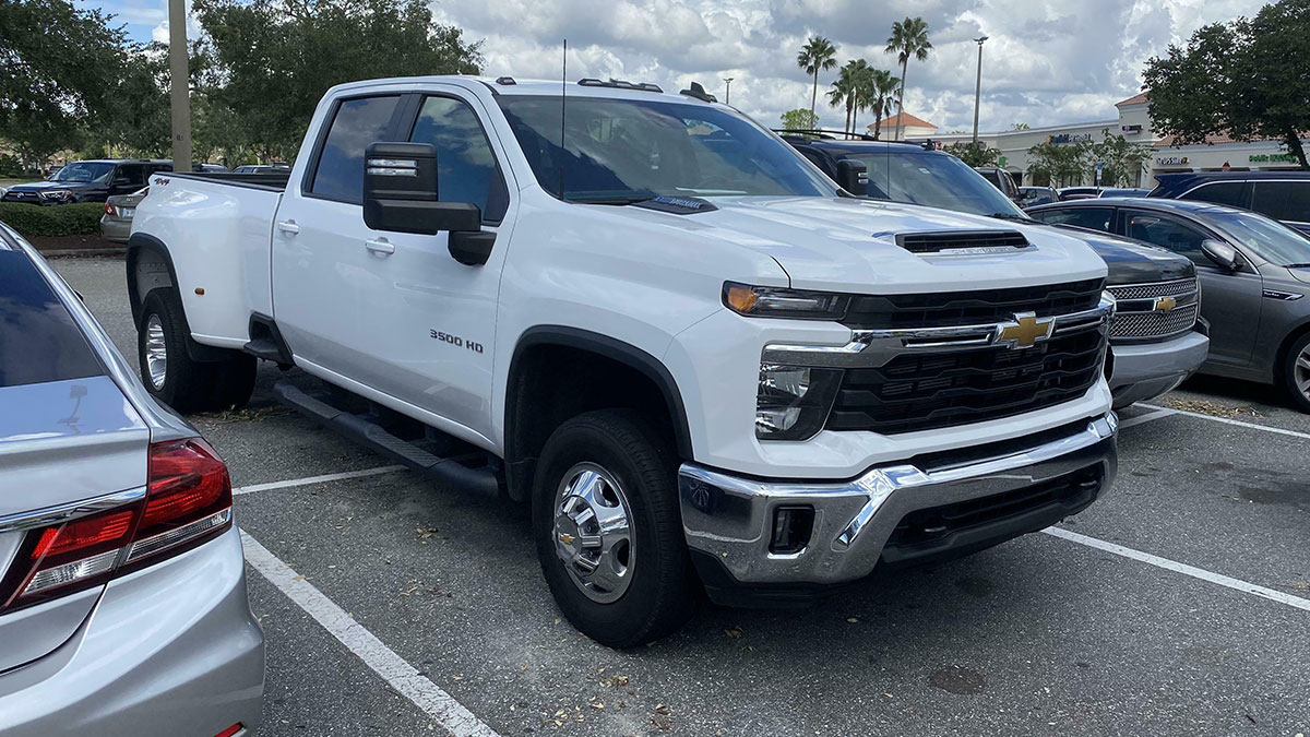 White Chevrolet 3500 HD pickup truck parked in a crowded lot under a cloudy sky with palm trees nearby.